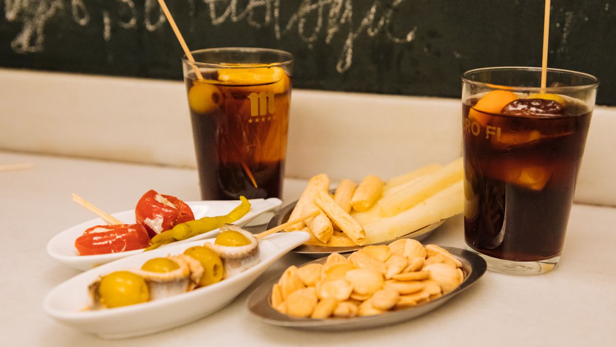 On a bar, two vermouth cocktails are flanked with dishes of snacks including fries, marcona almonds, and anchovy-swathed olives.