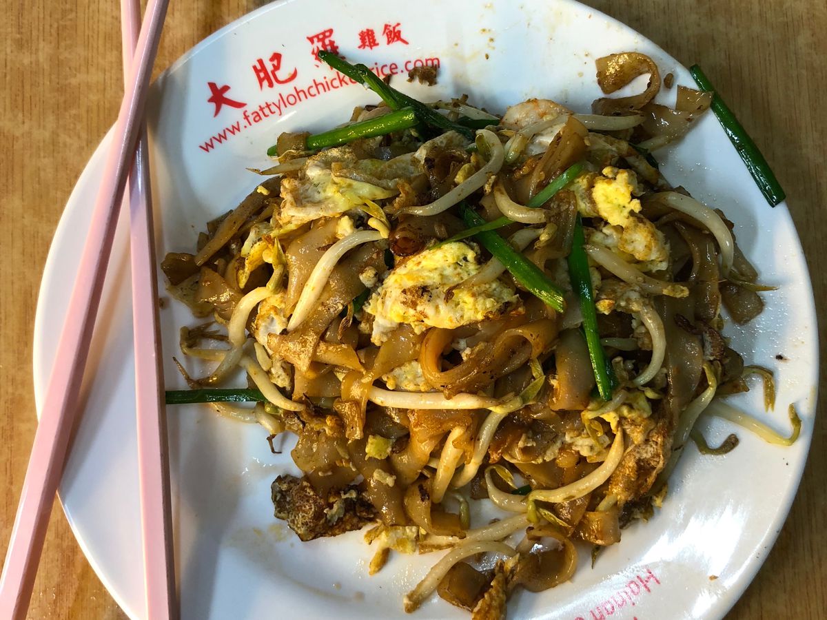 From above, a plate of fried rice noodles with bean sprouts, greens and onions, with a pair of chopsticks balanced on the rim on a wooden tabletop