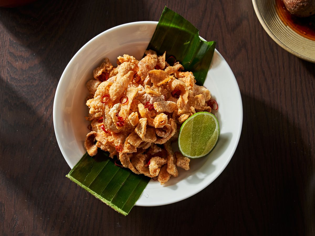 An overhead shot of a pile of crispy chicken skin strips set on a white plate with a green banana leaf laid down the middle. A lime wedge is on the side of the plate.