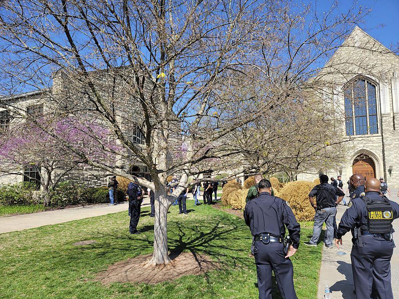 Blue uniformed officers mill about a green lawn with bare trees. Gothic inspired buildings can be seen in the background.