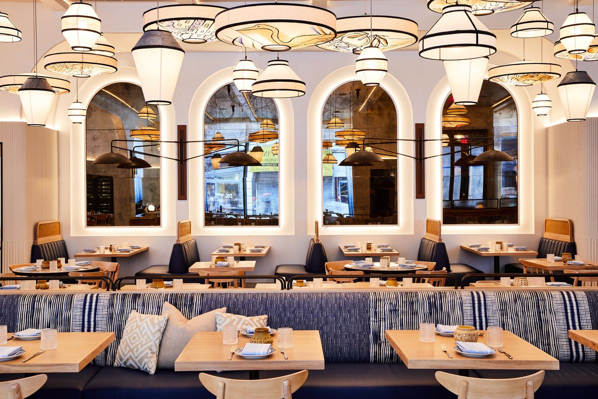 A light and warm restaurant interior with blue banquettes, arched windows, and neutral-toned pendant chandeliers.