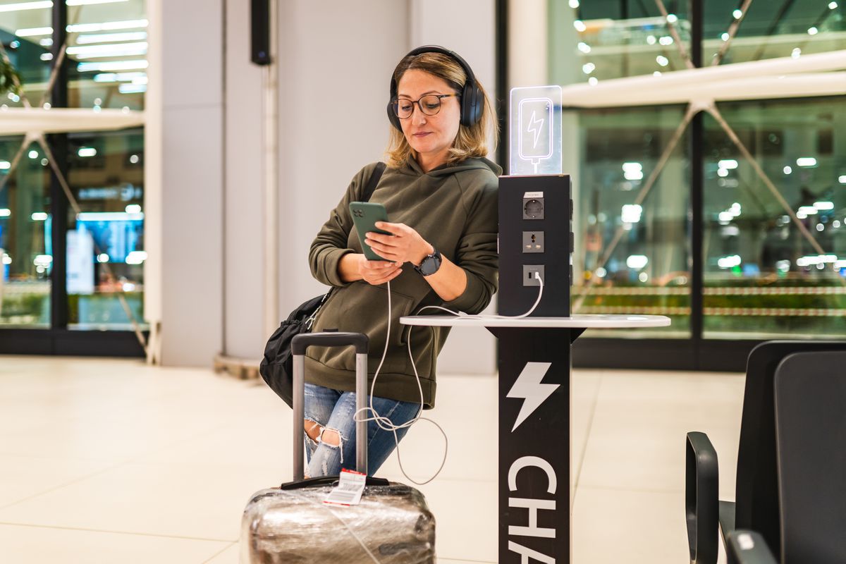 A woman charging her phone at the airport. She wears an army green sweatshirt and large black headphones.
