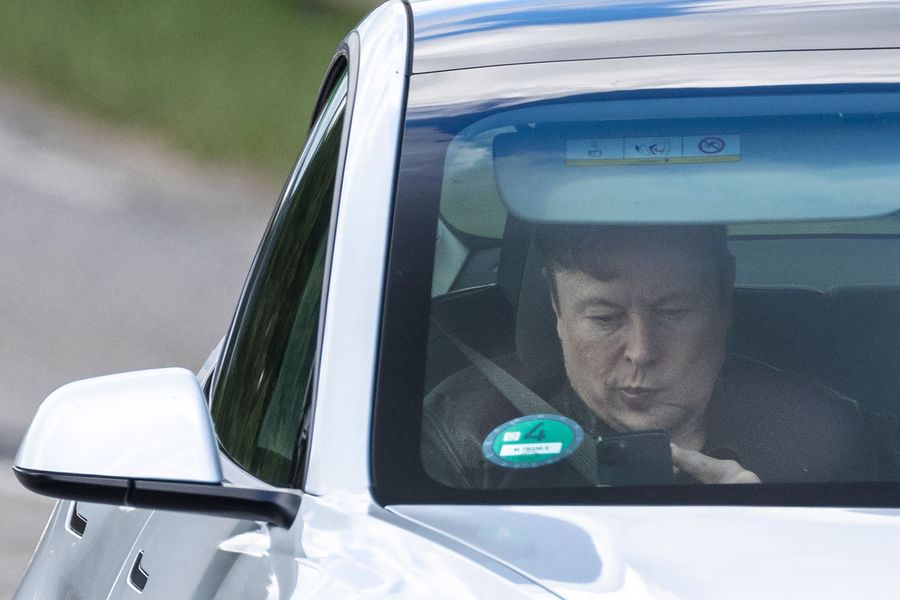 Elon Musk looks at his phone while sitting in the passenger seat of a car.