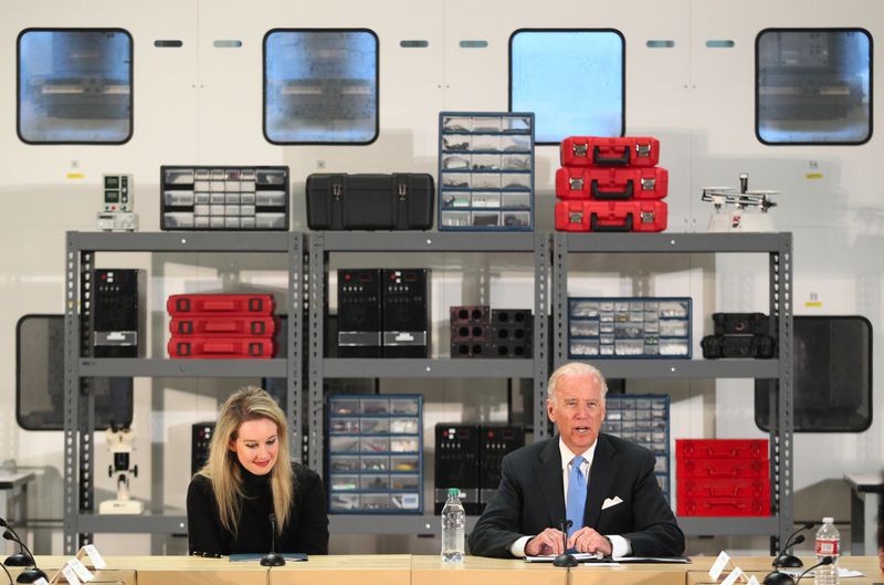 Elizabeth Holmes sitting next to Joe Biden in front of shelves of scientific-looking hardware.