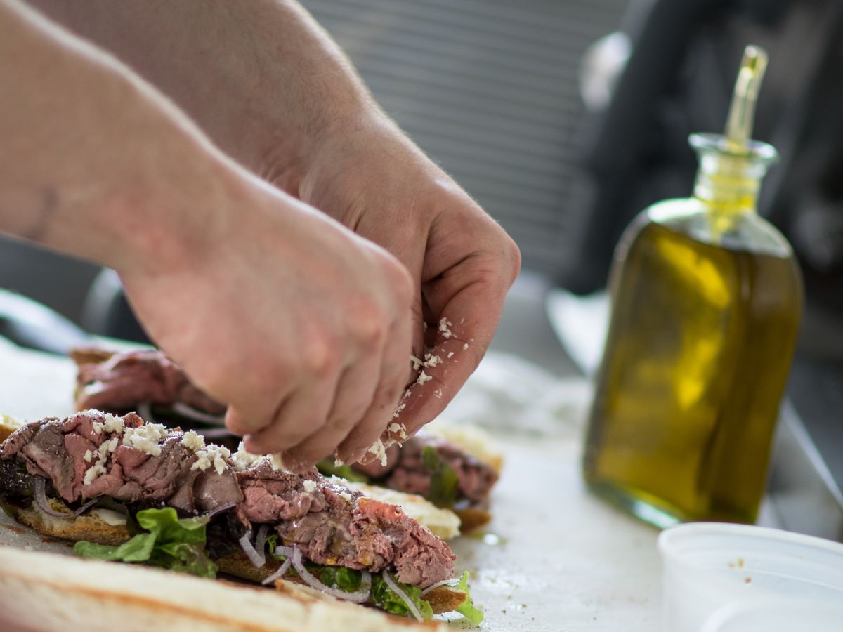 Two hands place shaved beef, lettuce and cheese on a baguette. A bottle of olive oil is visible in the background.