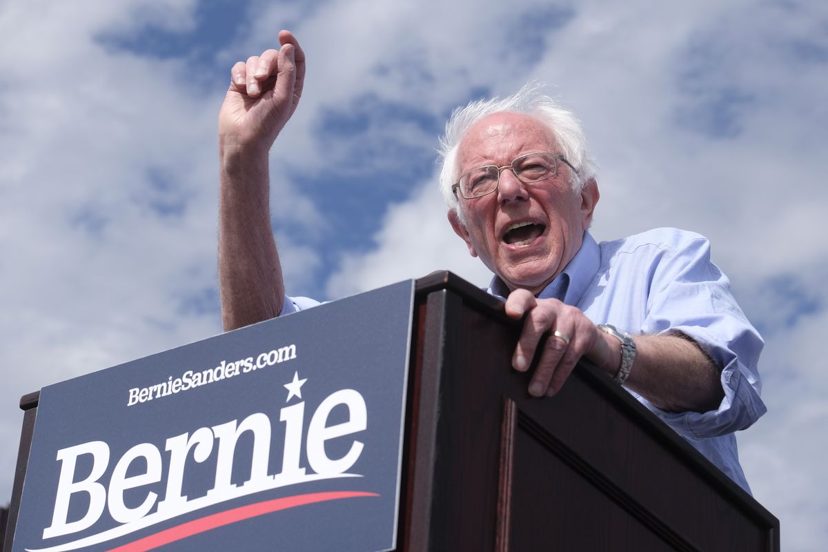 Sanders gestures, shouting, at a podium bearing his name under a blue sky dotted with white clouds.