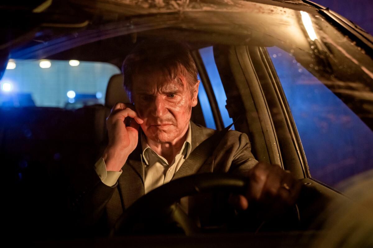 Liam Neeson, visibly disheveled, sits behind the wheel of a car with a cell phone next to his ear in Retribution.