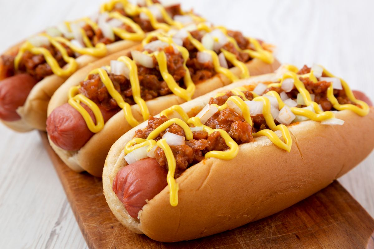 Three hot dogs on buns topped with brown chili, diced onion, and yellow mustard.