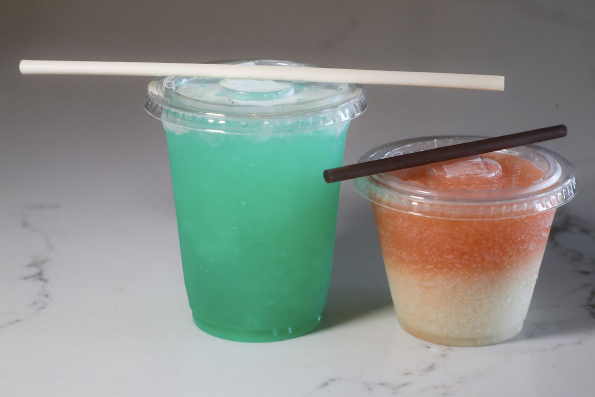 Two colorful takeout cocktails are placed on a granite countertop with sealed lids and plastic straws.