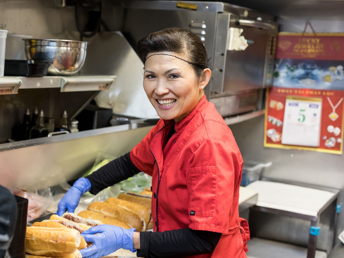 A smiling woman in a red shirt and hair net in the kitchen at Quang, building banh mi