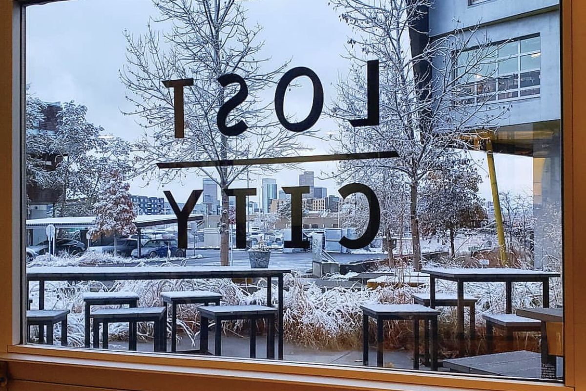 A photo taken out the window of Lost City coffee showing Lost City coffee lettering and a view of the patio with downtown Denver in the distance 