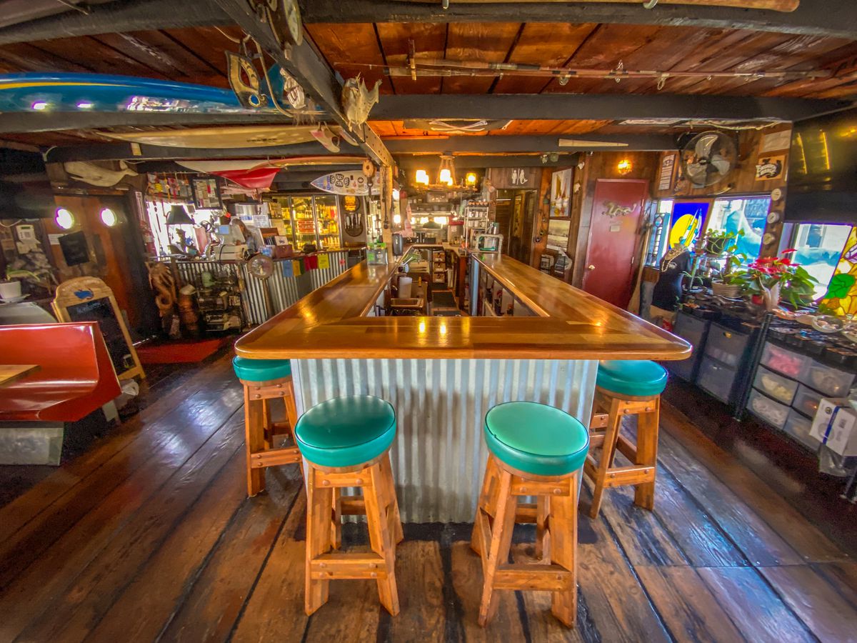 Teal-colored bar stools wrap around a small wooden bar inside a restaurant at daytime.
