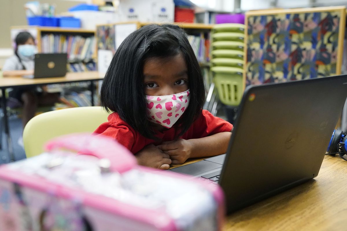 Young girl in a mask printed with pink hearts, works on her laptop at a learning center.