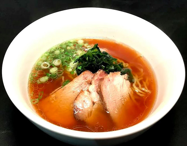 A dark brown broth with three pieces of pork, chopped green onions, and noodles in a white bowl