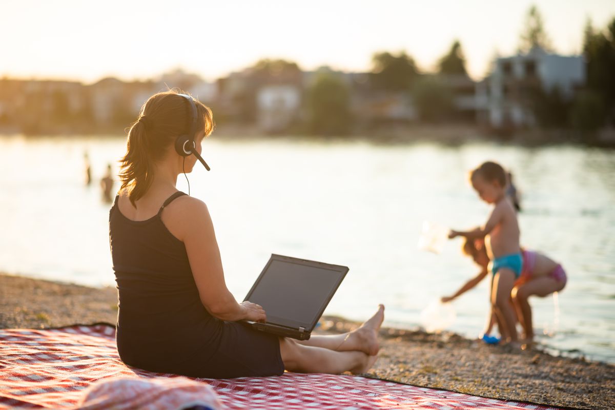 A woman sits on a blanket at the shore while working on her laptop computer and watching her children play near the water.
