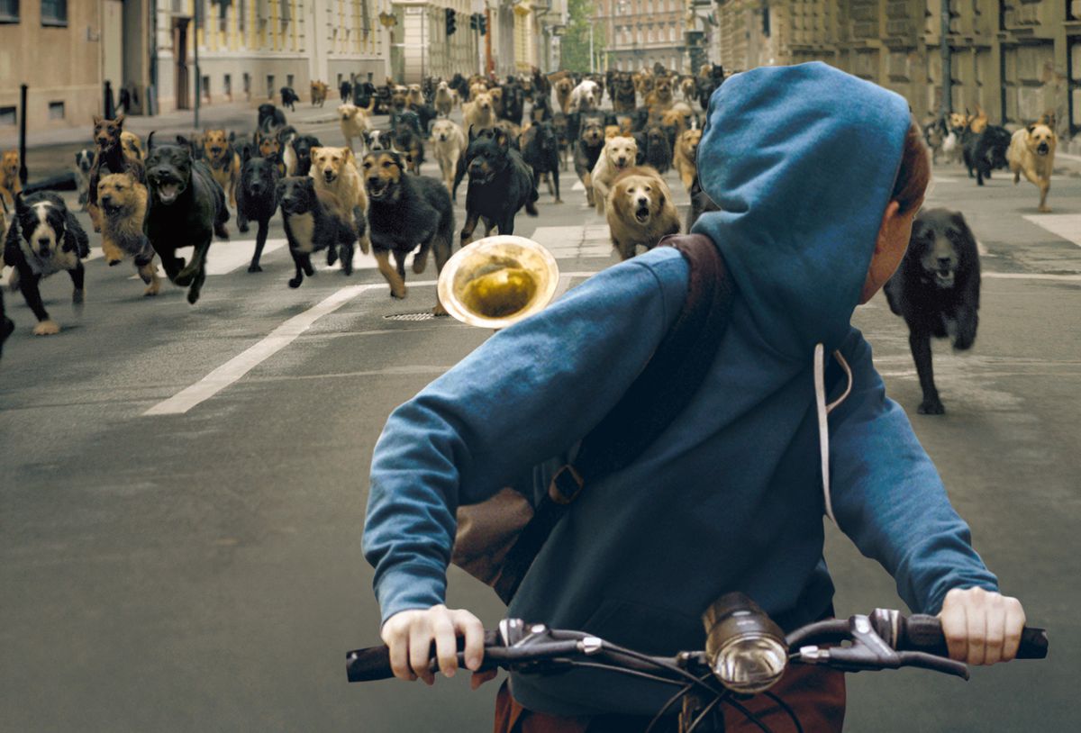 A girl (Zsófia Psotta) in a blue hoodie riding a bike with a brass trumpet in her backpack looks back at a mass of dogs racing down an empty street.