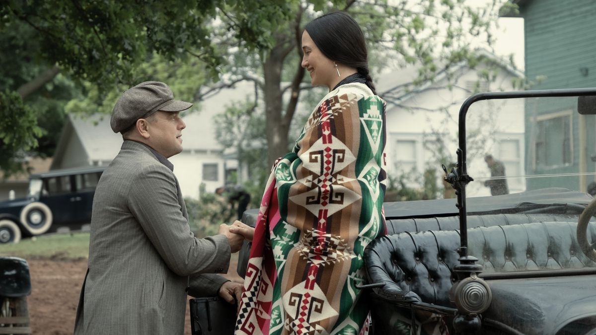 Leonardo DiCaprio, in a chauffeur’s cap, helps Lily Gladstone, wearing a colorful blanket, down from an old-timey car in Killers of the Flower Moon