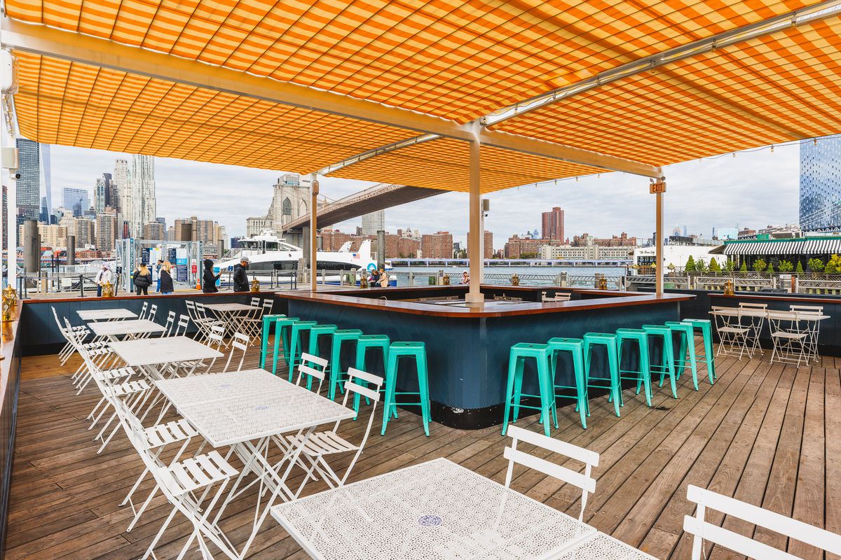 White tables and bar stools make up an outdoor bar along a waterfront in Brooklyn’s Dumbo neighborhood.