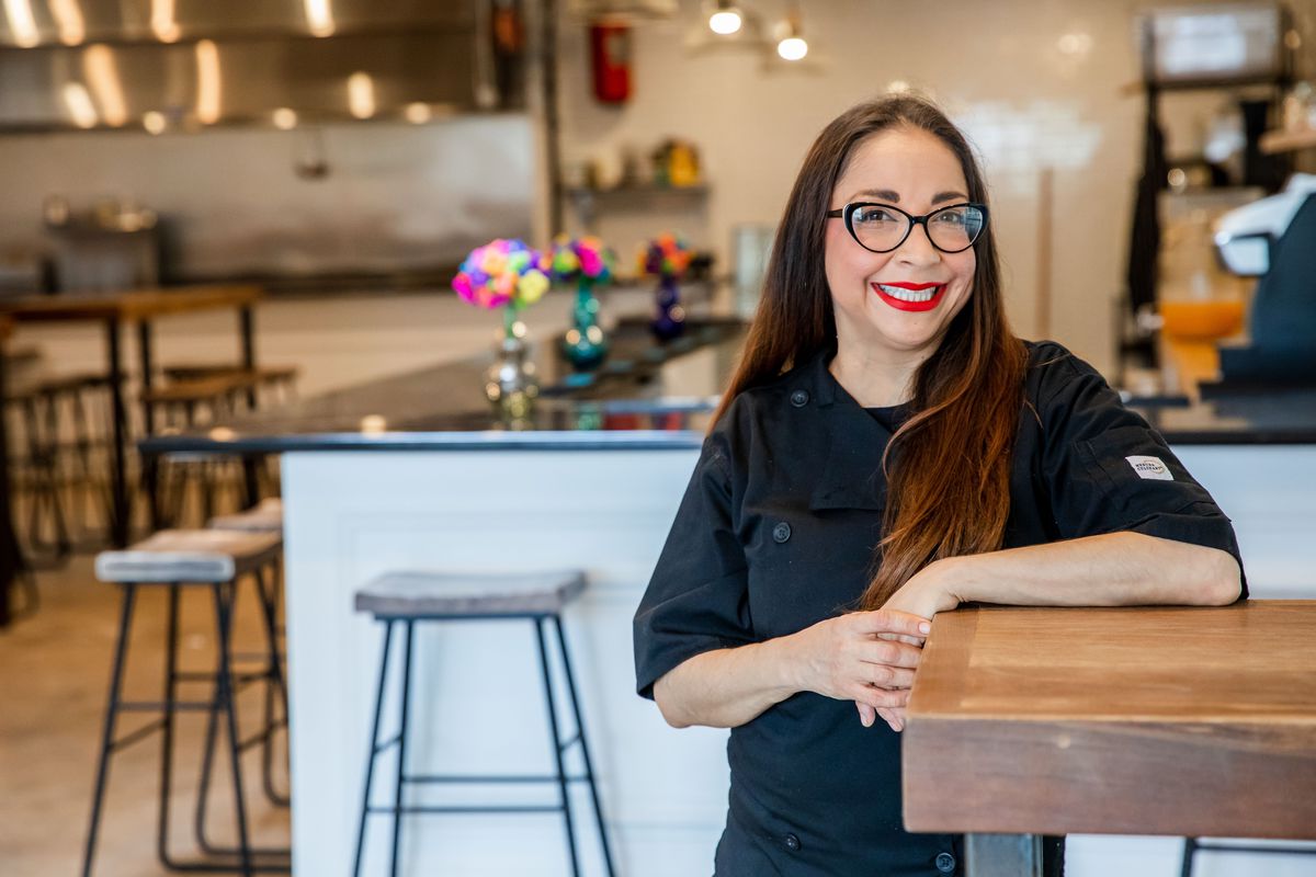 a chef in a black kitchen jacket, with long black hair and red lipstick, leans against the bar at her newly-opened Tex Mex restaurant