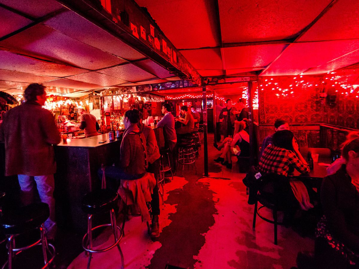 An underground-looking room with a bar on the left is dimly lit by red-tinted light and string lights. People stand around the bar and sit in parallel tables to the right.