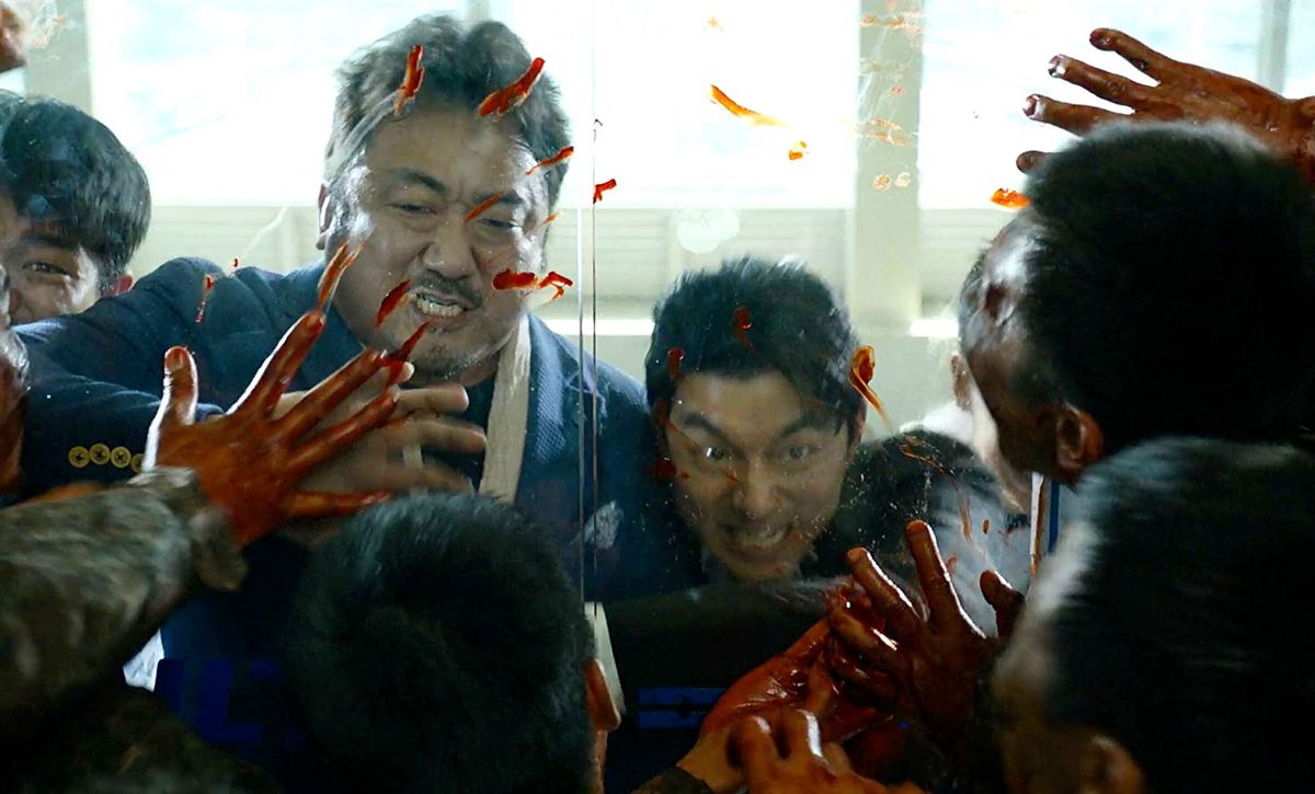Two men straining to hold a pair of glass doors shut as a horde of zombies barrage them, blood stains visible on the glass surface.