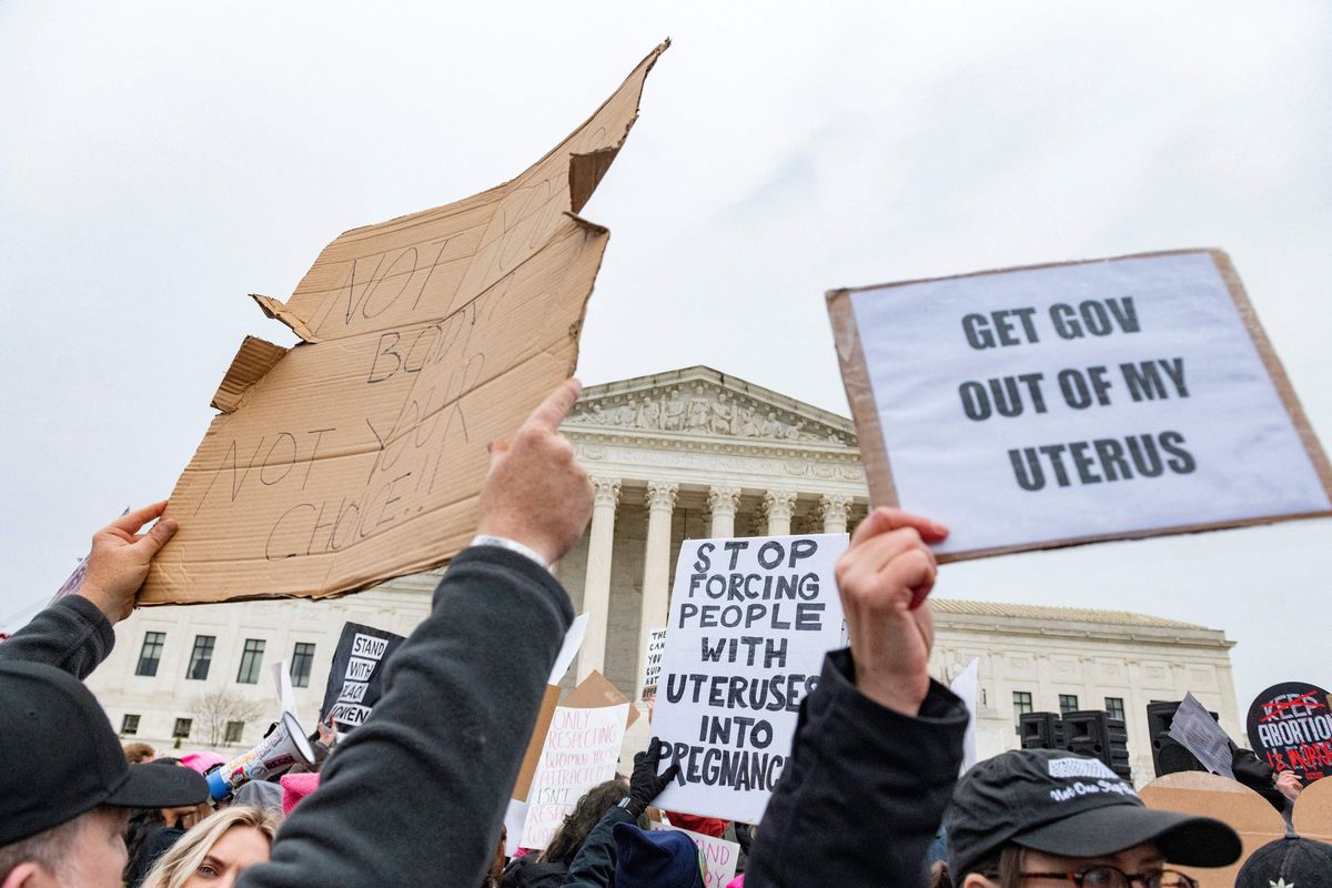 A crowd of people, seen from head-height, some holding signs in the air made from cardboard and paper with various slogans related to abortion, all in front of the white-pillared Supreme Court building.