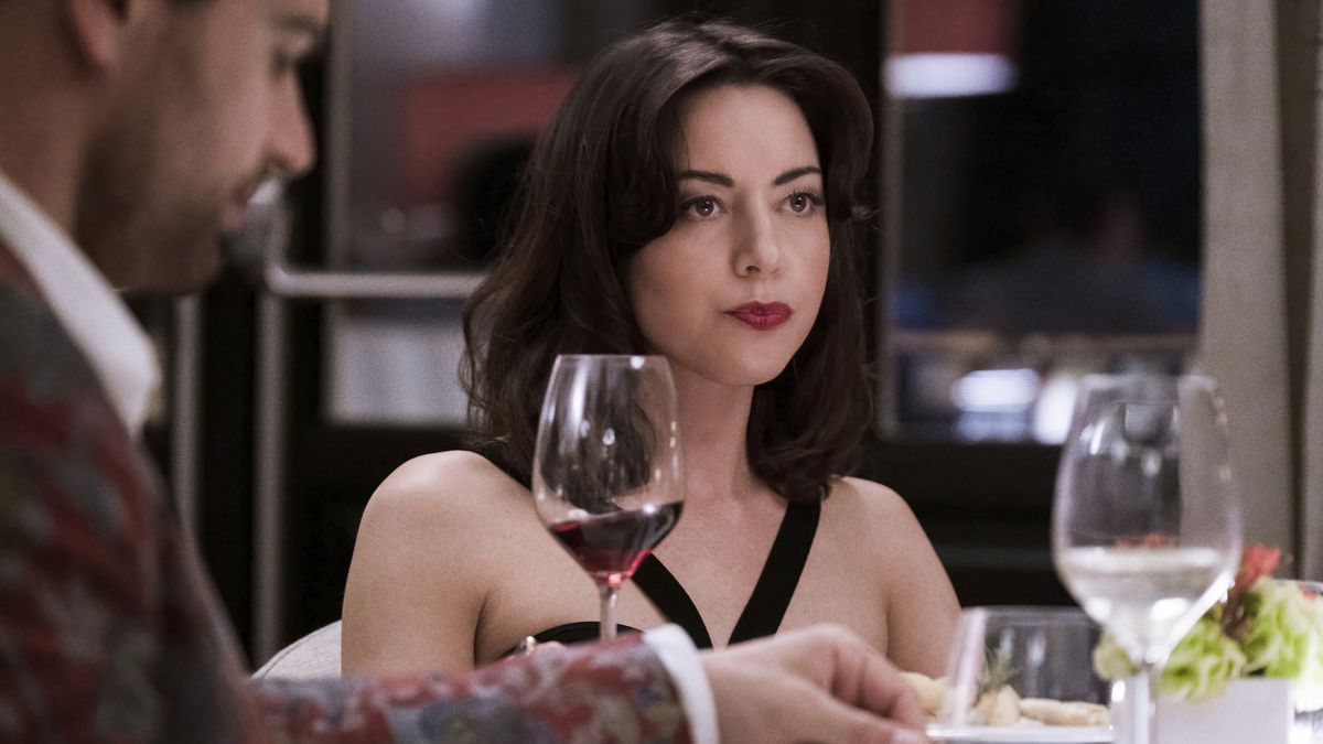 A young woman in a black dress looks across a fancy dinner table with pursed lips.