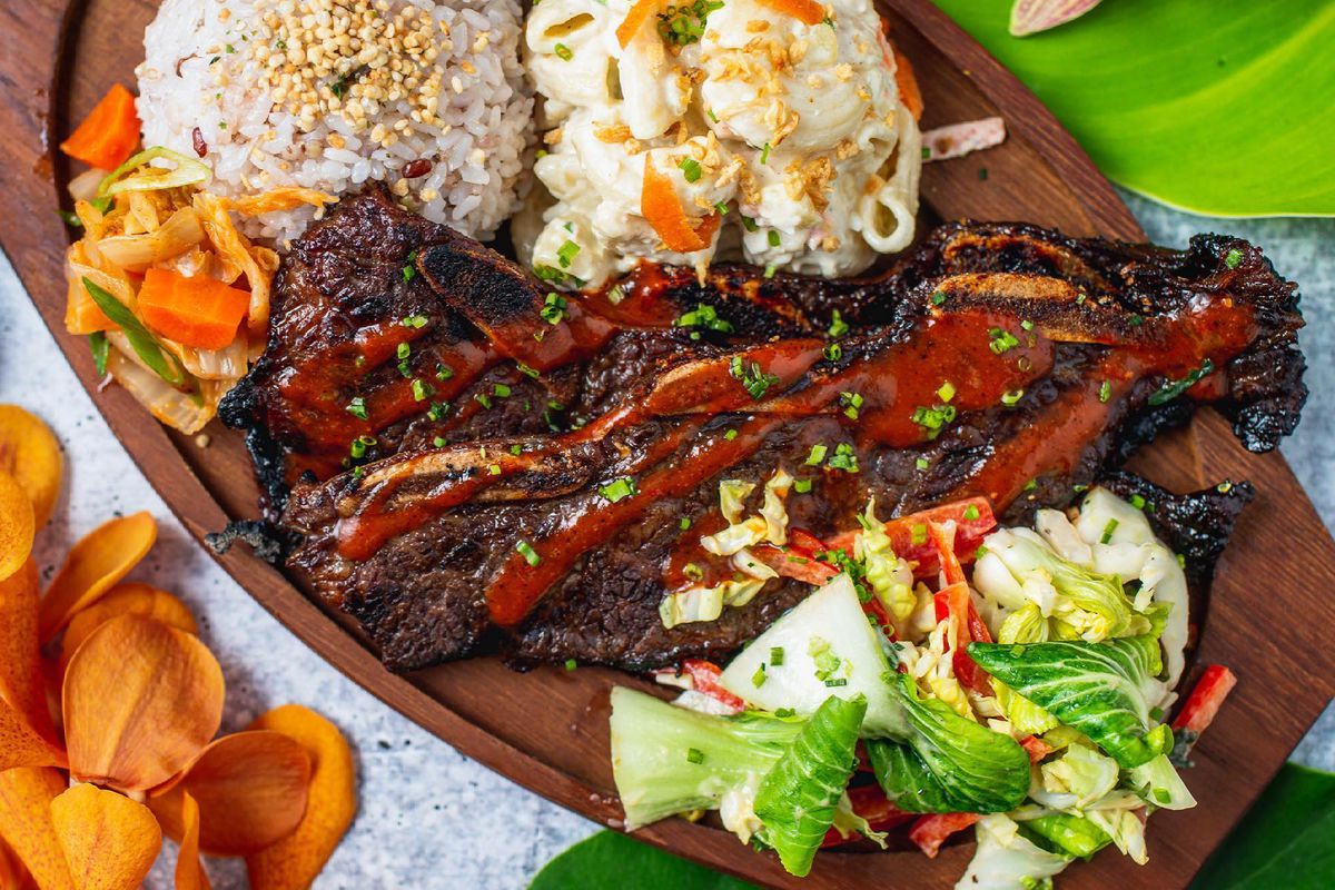 A wooden platter holds macaroni salad, rice, and short ribs with a spicy sauce at Da Pine Grinds