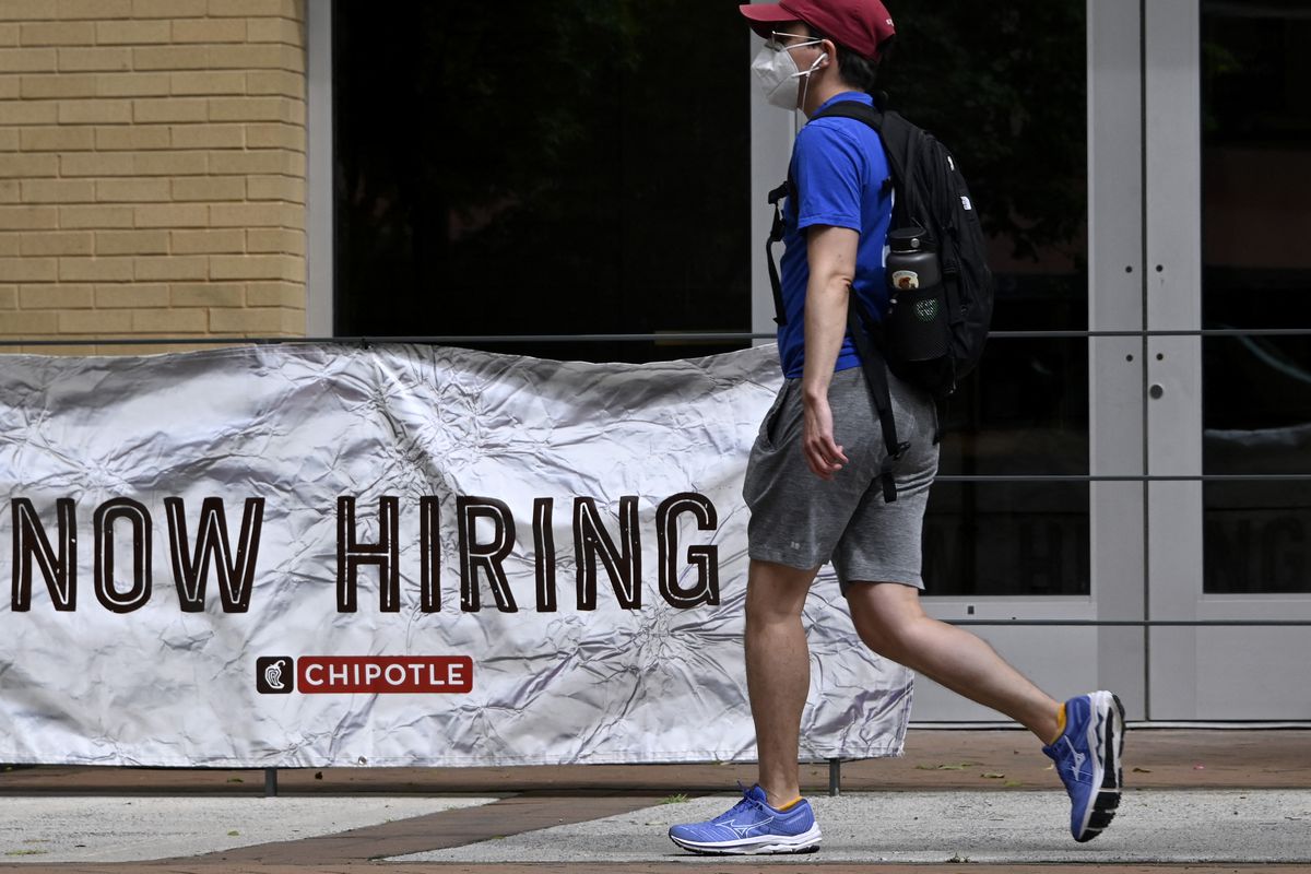 A man in a blue shirt and gray shorts walks past a restaurant with a large “Now Hiring” banner outside.