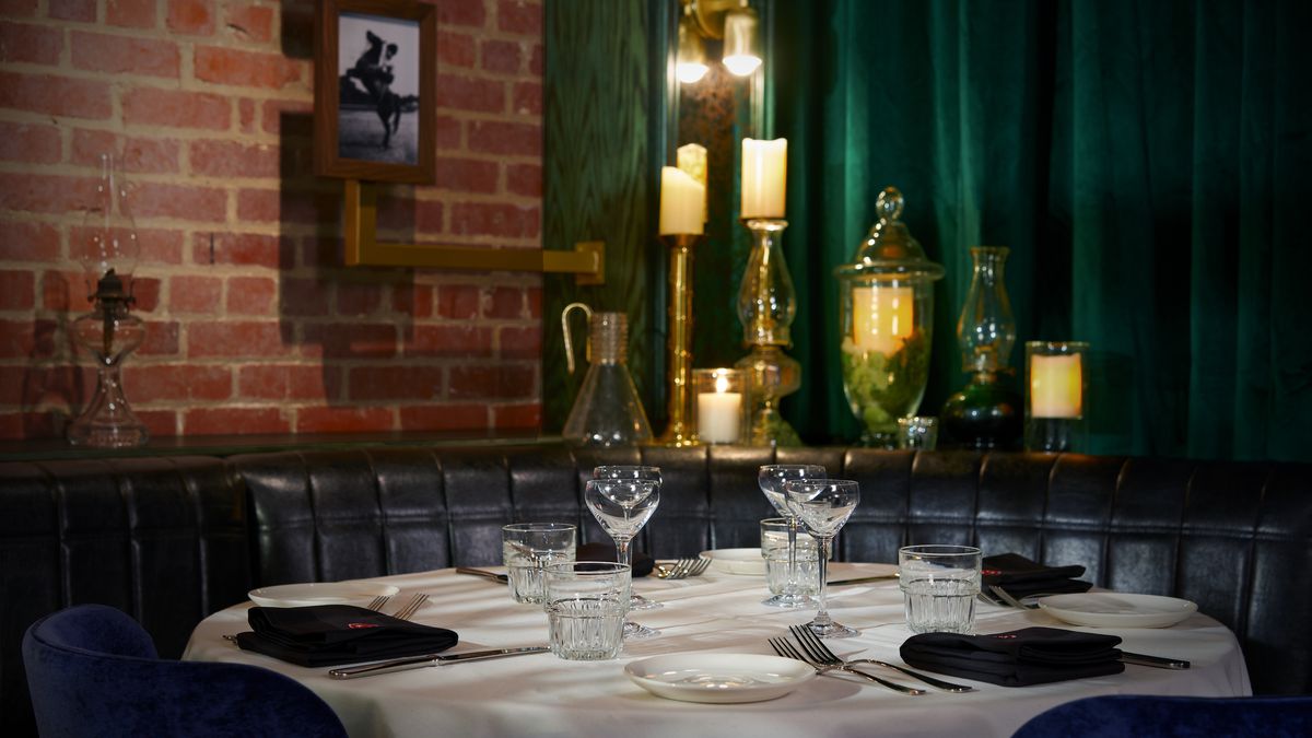A booth table is set with a white tablecloth and china, with green velvet curtains in back and candles lit on the table.
