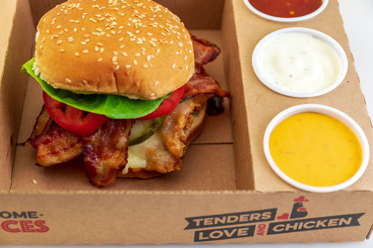 A photo of a chicken sandwich with a brown bun with lettuce, tomato, bacon, and chicken visible below it with ketchup, ranch and mustard cups ton the side