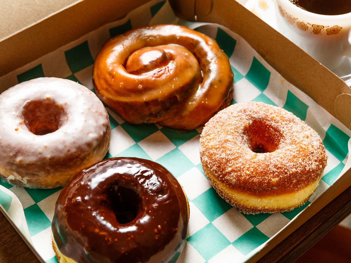 Three doughnuts, one of them covered in chocolate, and a tamarind honey bun sit on a seafoam green and white checkered sheet of paper in an open doughnut box.