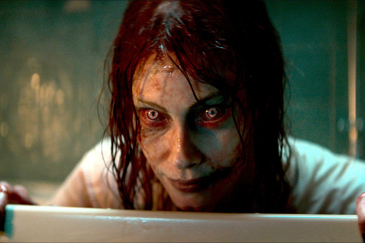 Ellie (Alyssa Sutherland), possessed and turned into a red-and-yellow-eyed, greasy-haired, grimy-faced Deadite, smiles eerily over a barrier in Evil Dead Rise