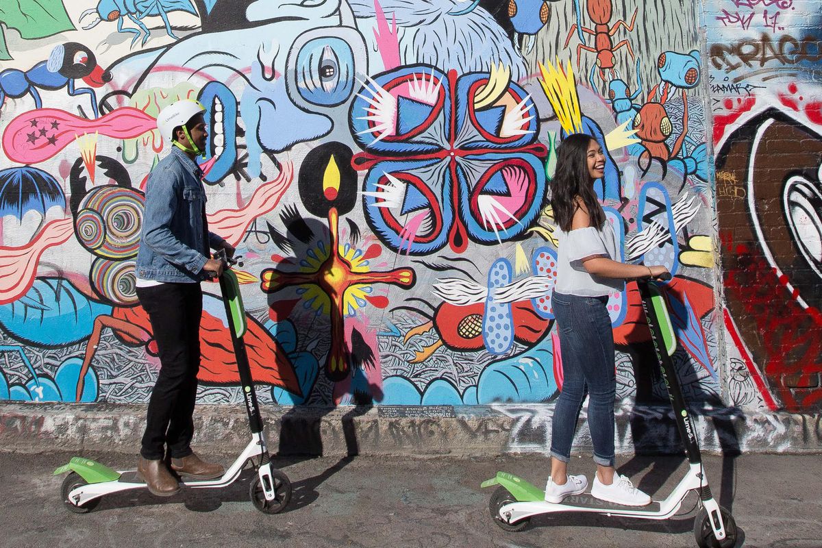 A picture of two people using lime scooters past a heavily decorated wall.