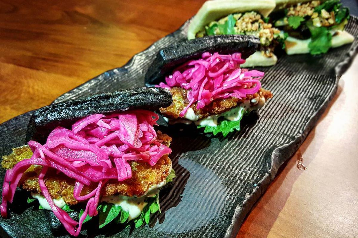 Four bao are on a long, narrow tray. The two in the forefront are black and made of squid ink. Bright pink pickled onions are visible inside.