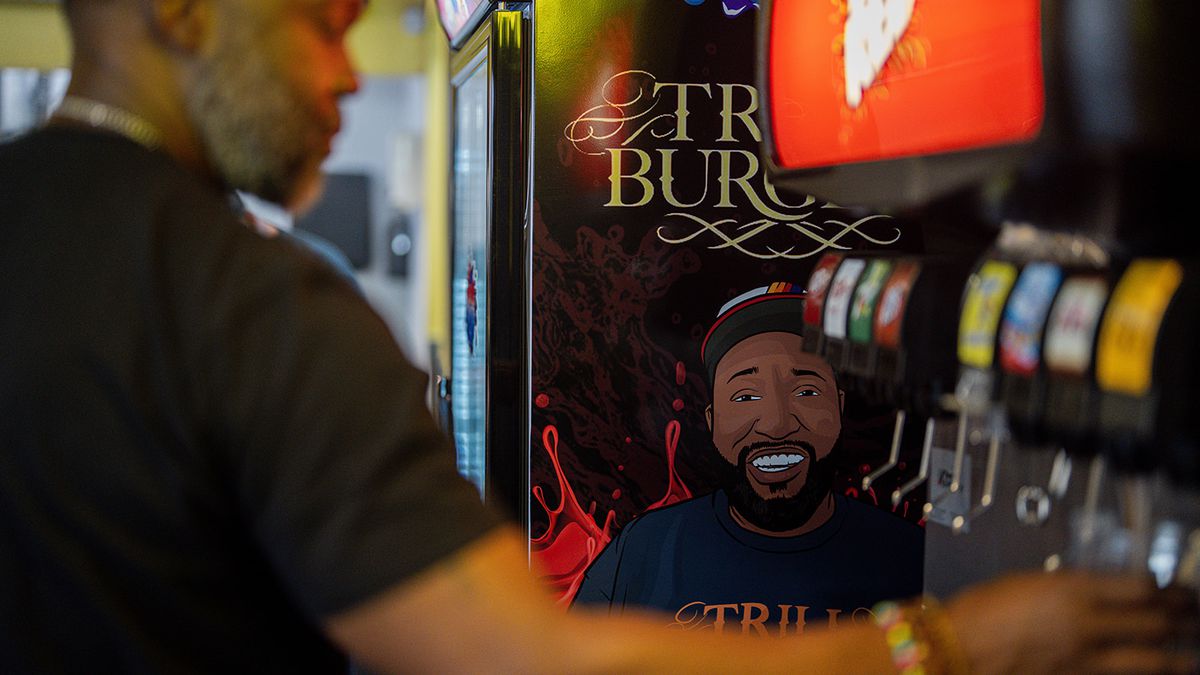 A man gets a drink from Trill Burgers’ soda fountain. Nearby, an illustration of a smiling Bun B.