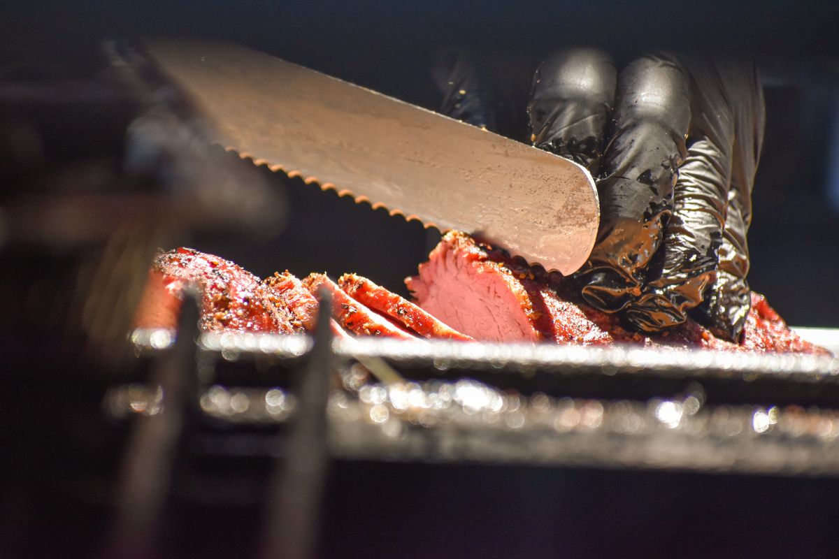Knife slicing through smoked meat, hands wearing black gloves.