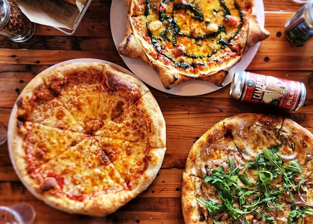 An overhead shot of small round pizzas on a wooden table with beers nearby.