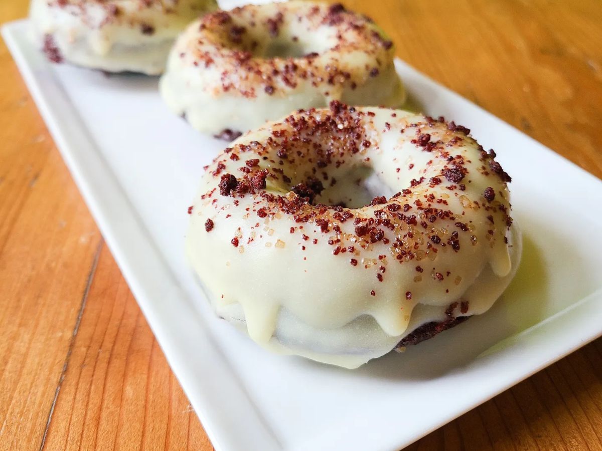 Three red velvet doughnuts sit on a rectangular white platter on a wooden table. The doughnuts have a white icing on top with dark red sprinkles on top.