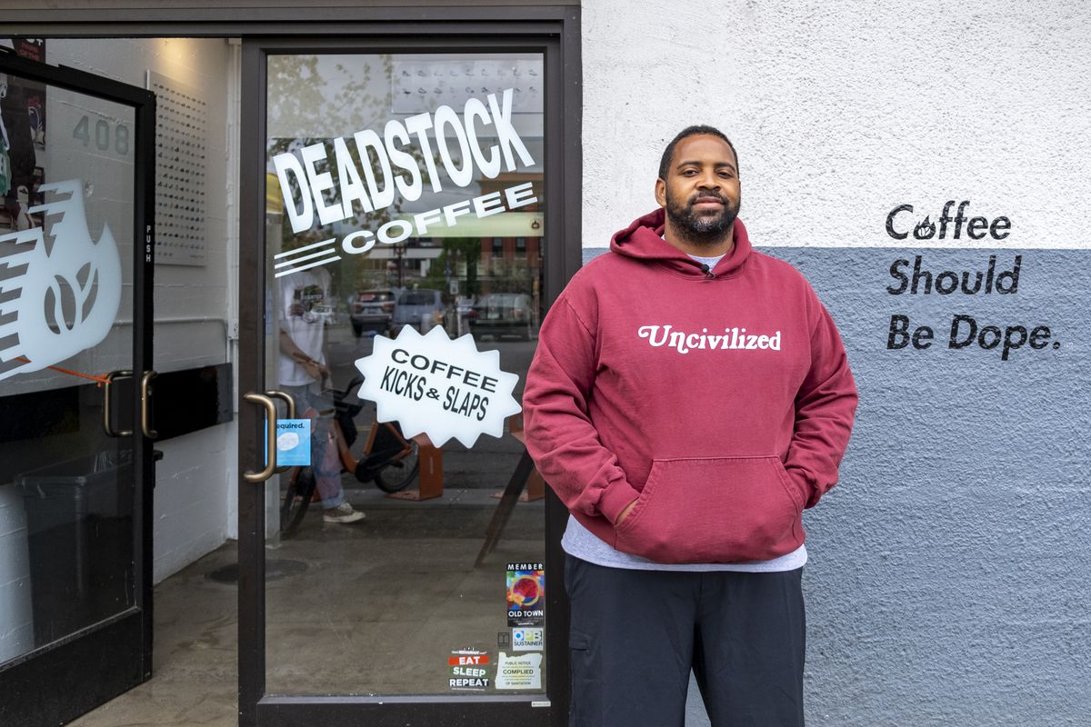 A man in a red sweatshirt that says “Uncivilized” on it stands outside a white-and-blue cafe. On the wall, the words “Coffee should be dope” can be read.