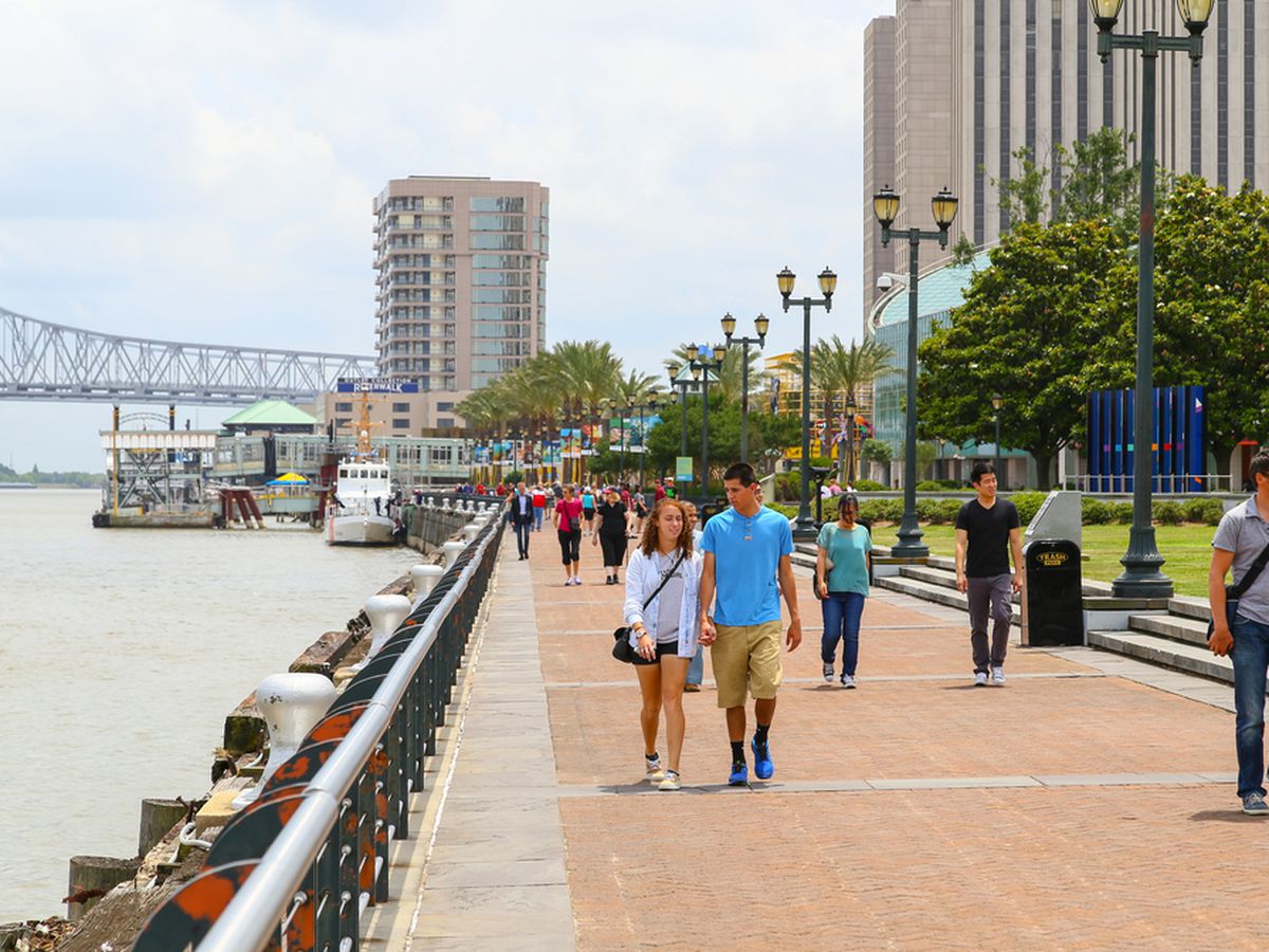 An outdoor scene of people walking down a sidewalk with the Mississippi River to the left and the Crescent City Connection and high rises in the back