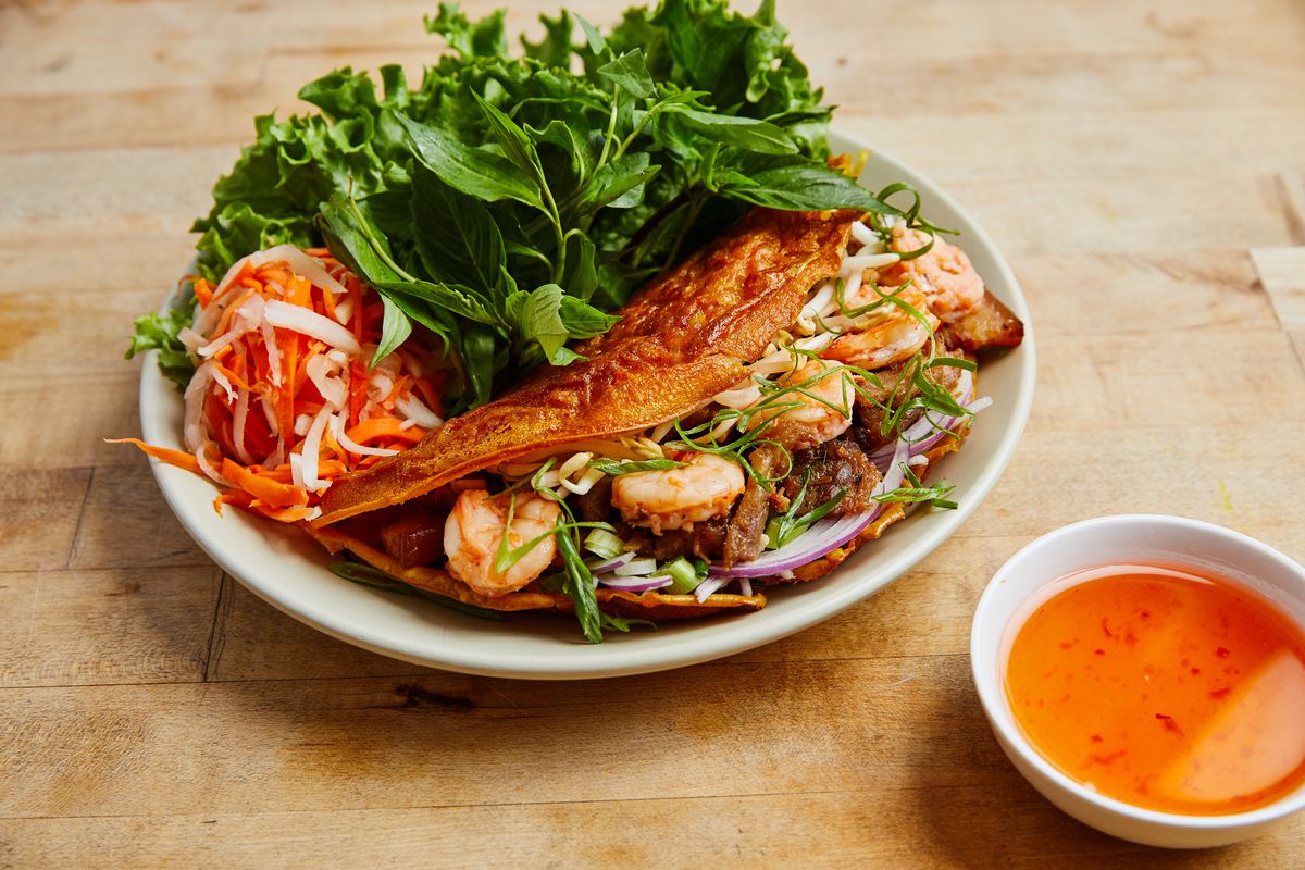 A white plate with green leafy lettuce, pickled strips of carrot and onion, and a fried pancake stuffed with shrimp, pork, and vegetables. A white cup of fish sauce for dipping is set off to the lower right side. The plates are set on a light wooden table.