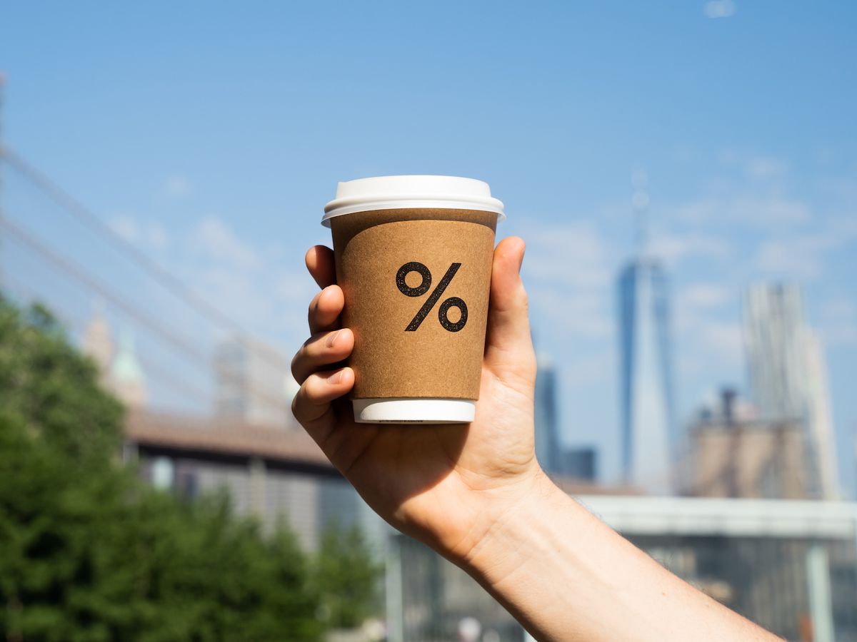A hand holding a brown coffee cup emblazoned with the % Arabica logo, against a backdrop of the Brooklyn Bridge