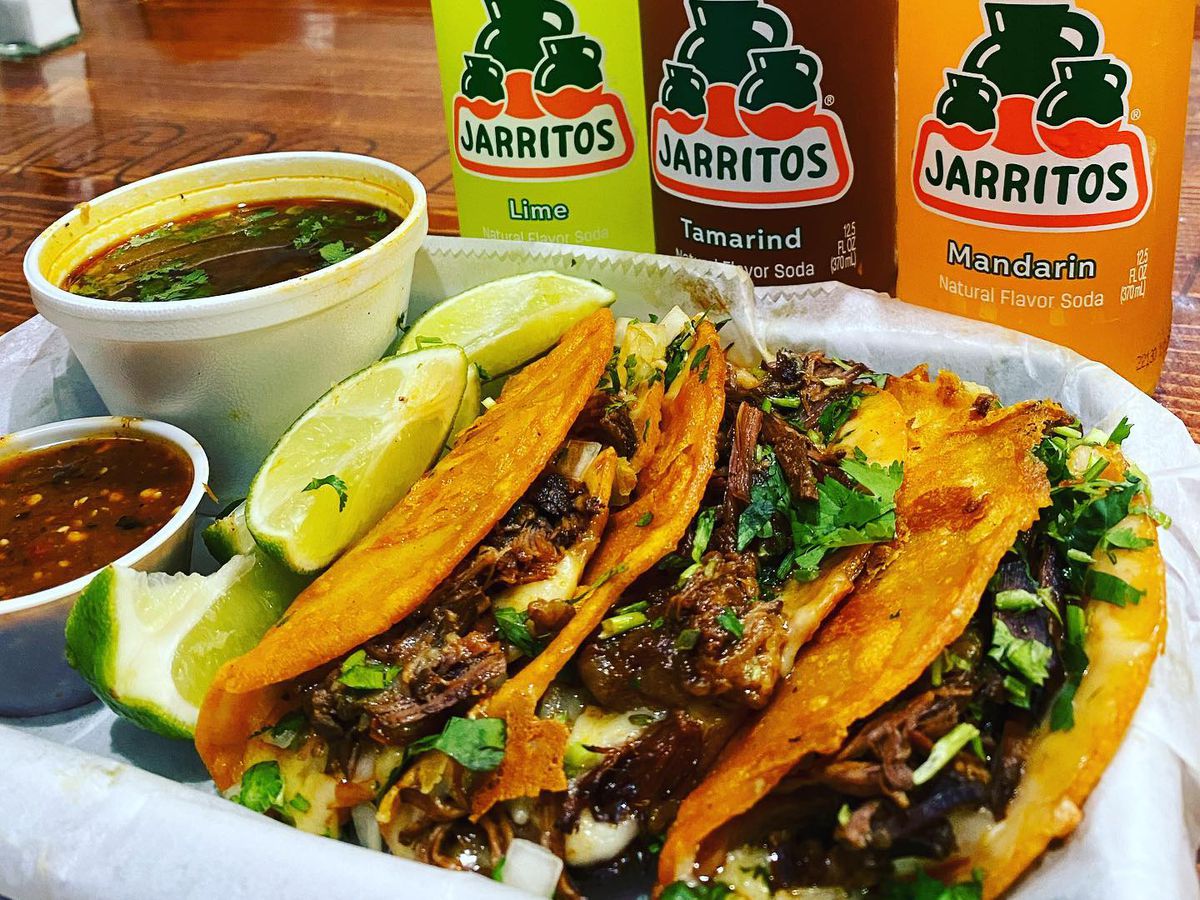 Three birria tacos dressed with cilantro and onions sit in a basket beside cups of consomme (birria broth) and three bottles of lime, tamarind, and mandarin orange Jarritos soda from Tacos La Villa in Smyrna, GA.