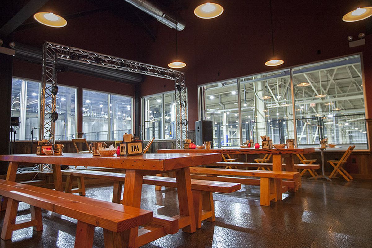 A large taproom space with long wooden tables and benches. Brewing equipment is visible through large windows.