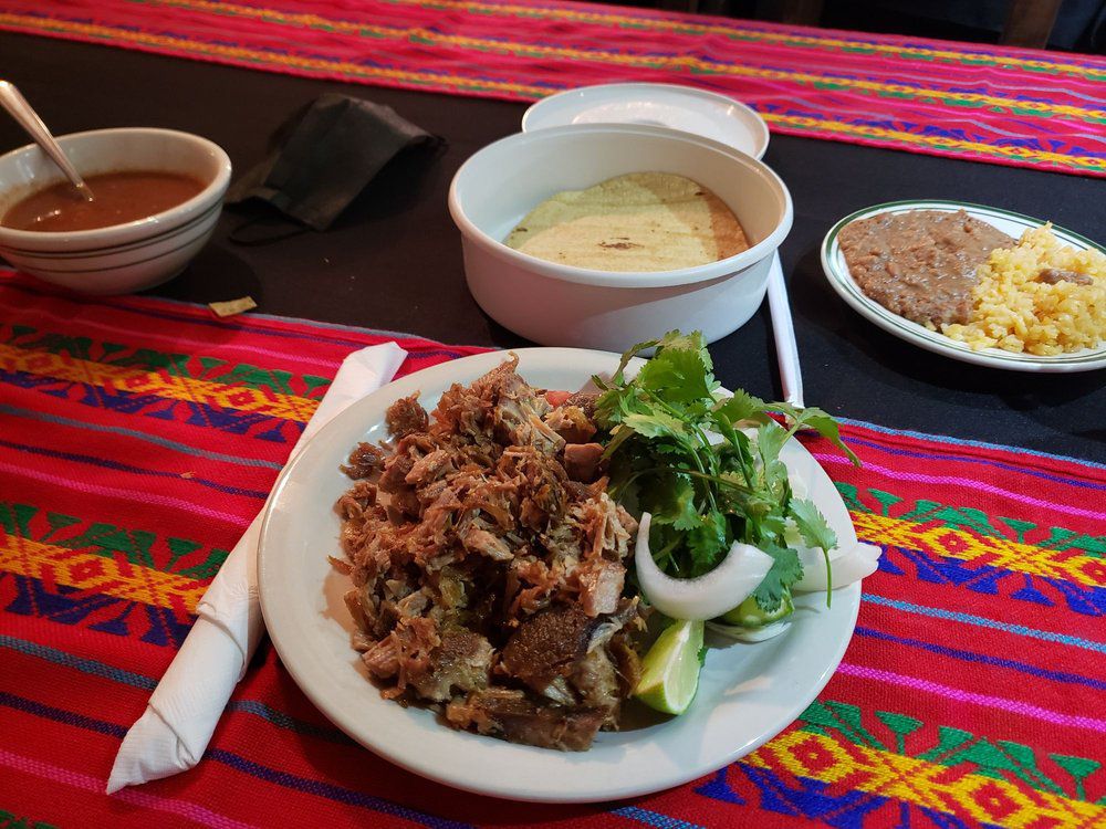 Carnitas, tortillas, rice, beans, salsa and cilantro served in white plates and bowls over a colorful Mexican textile tablecloth. 