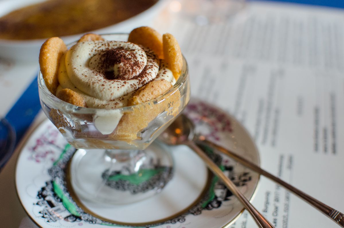 A swirl of tiramisu is in a glass stemmed bowl sitting on a decorative vintage plate. Two long spoons accompany it.