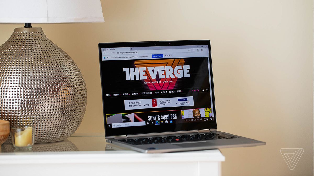 The Lenovo ThinkPad X1 Titanium Yoga open on a bedside table next to a lamp, angled to the right. The screen displays The Verge’s homepage.