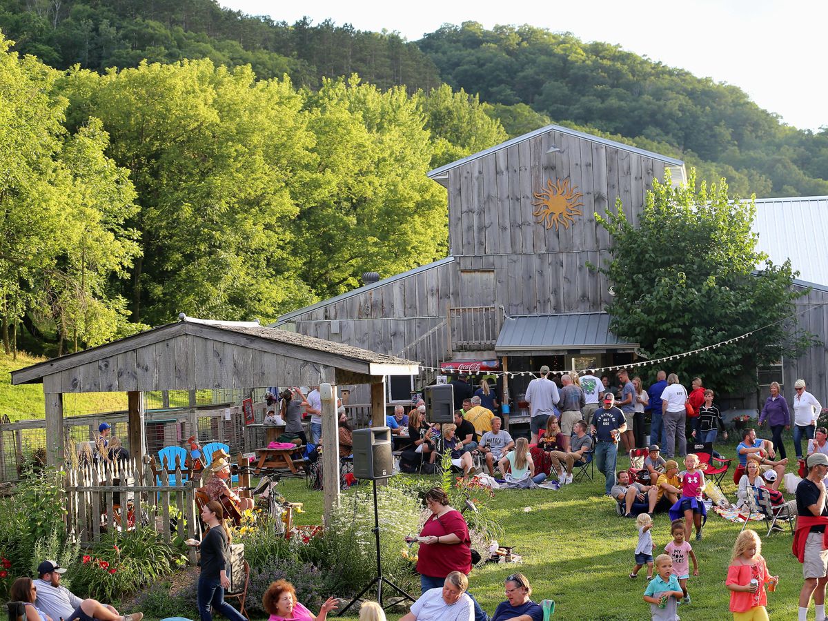 A large wooden barn with many people on the grass lawn in front of it. 