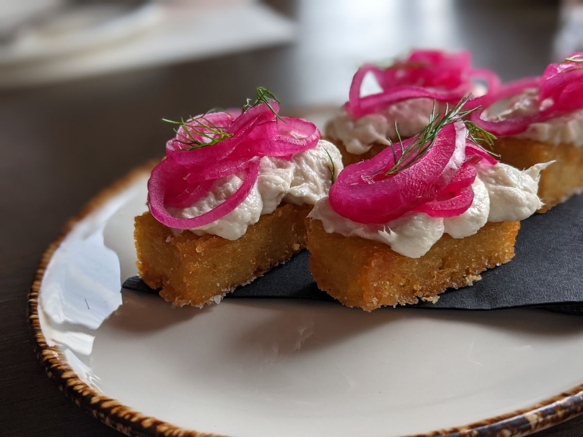 Four large fried tater tots on a plate, topped with fluffy piles of white fish and pink pickled onions